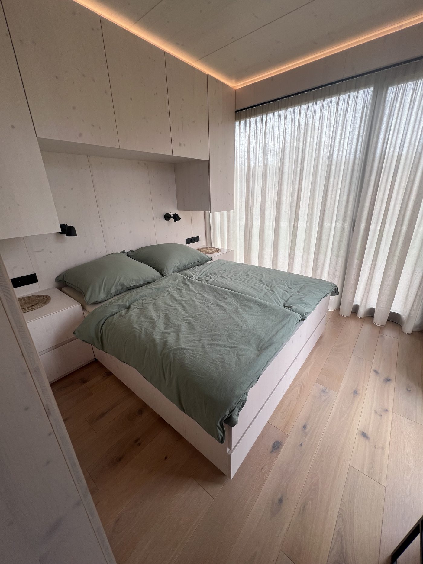 Bedroom with sage-green bed linen, built-in wardrobes with indirect lighting and sheer curtains
